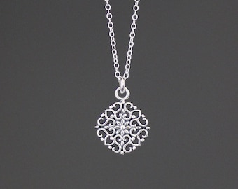 Può includere: Una collana d'argento con un delicato ciondolo ornato con un designo circolare con intricati vortici e un piccolo fiore al centro.