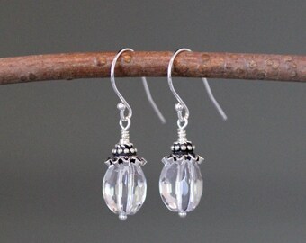 Puede incluir: Un par de pendientes de cristal transparente y facetado con detalles metálicos plateados. Los pendientes tienen forma de lágrima y cuelgan de ganchos plateados. El diseño presenta una tapa con cuentas y una pequeña cuenta plateada en la parte inferior.