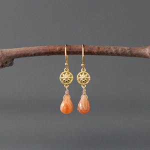 May include: A pair of gold drop earrings featuring a circular floral charm connected to a faceted, teardrop-shaped orange gemstone. The earrings hang from gold hooks, showcasing a delicate metal design with warm, vibrant stone accents.