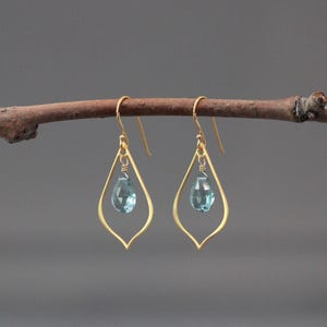 Puede incluir: Un par de pendientes colgantes dorados con una gema azul en forma de lágrima suspendida dentro de un delicado marco calado. Los pendientes se exhiben en una rama natural sobre un fondo gris neutro.