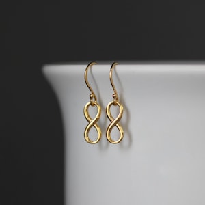 May include: A pair of gold infinity symbol earrings with a delicate design. The earrings are hanging from a white surface.