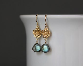 May include: Gold-toned dangle earrings. Each earring features a floral design at the top, with a teardrop-shaped, faceted, iridescent gemstone pendant. The earrings are displayed against a white background.