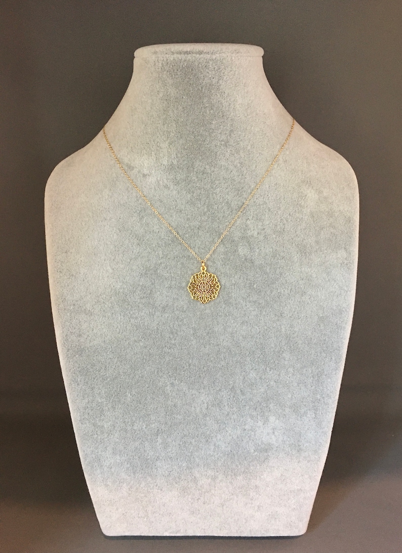 May include: A gold necklace with a delicate, intricate pendant in the shape of a mandala. The necklace is displayed on a gray velvet bust form.
