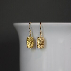 May include: A pair of gold-colored earrings with a rectangular shape and intricate, swirling patterns. The earrings hang from delicate gold hooks and feature a small bead at the bottom. The jewelry is displayed against a white background.