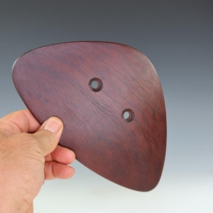 May include: A dark reddish-brown, teardrop-shaped wooden object with two evenly spaced holes. The wood grain is visible, and the object is held in a person's hand.