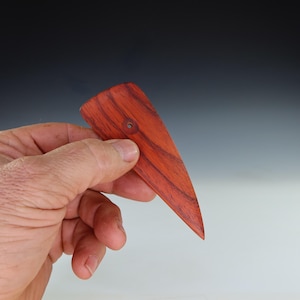 May include: A handcrafted, reddish-brown wooden tool with a triangular shape and a small hole near the top. The wood grain is visible, and the tool is held in a hand against a blurred background.