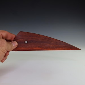 May include: A reddish-brown wooden pottery rib tool with a triangular shape and a small hole. The tool is held in a hand and is designed for shaping and smoothing clay in pottery projects.