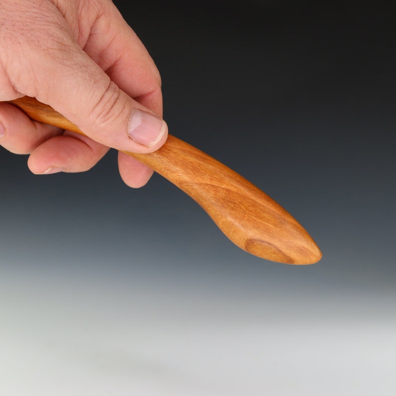 May include: A close-up of a wooden back scratcher with a curved handle and a rounded tip. The back scratcher is made of light brown wood with a natural grain.