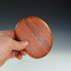 May include: A smooth, oval-shaped wooden coaster with a rich reddish-brown color. The grain of the wood is visible and adds to the natural beauty of the coaster.