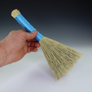 May include: A small broom with a handle wrapped in bright blue thread. The broom's bristles are made of natural straw. The broom is held in a hand against a white and gray gradient background.