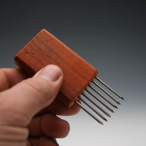 May include: A wooden tool with a rectangular handle and eight metal prongs. The tool is made of reddish brown wood and the prongs are silver.