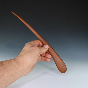 May include: A long, thin, wooden tool with a rounded tip. The tool is made of a reddish-brown wood and is held in a person's hand.