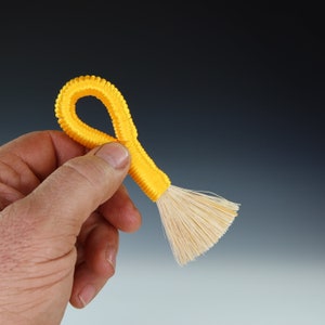 May include: A yellow, looped, string brush with a natural bristle head. The brush is held in a hand.