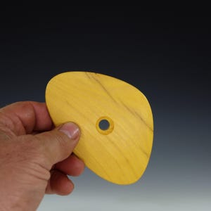 May include: A yellow wooden pottery rib tool with a hole in the center. The tool is held in a hand and has a smooth, curved shape. The wood grain is visible. The background is a gradient of dark blue to white.