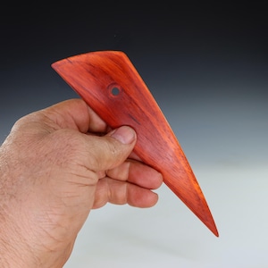 May include: A handcrafted, reddish-brown wooden pottery rib tool with a triangular shape and a small hole near the top. The tool is held in a hand, showcasing its smooth, polished surface and pointed end. This tool is used for shaping and smoothing clay.