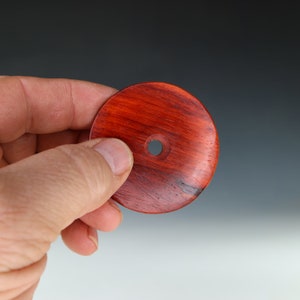 May include: A round, red wooden disc with a hole in the center. The disc has a natural wood grain pattern and a smooth, polished finish.