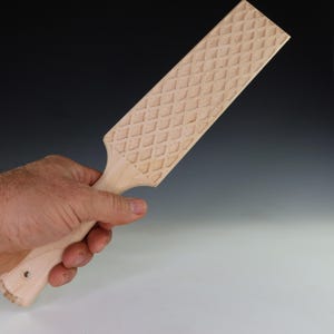 May include: A light-colored wooden paddle with a diamond pattern carved into the surface. The paddle has a handle with a hole at the end. The paddle is held in a hand.