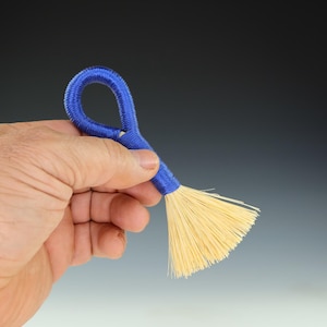 May include: A small blue brush with a handle made of braided cord. The brush has a fan-shaped head of natural bristles.