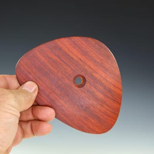 May include: A reddish-brown, teardrop-shaped wooden tool with a small hole near the center. The tool has a smooth, polished surface and is held by a hand. The wood grain is visible, adding texture to the design. This tool is likely used for pottery or woodworking.