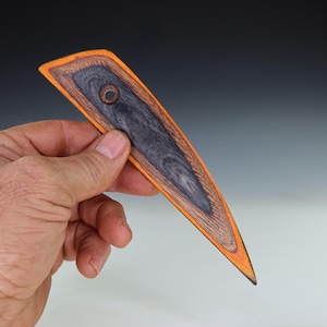 May include: A handmade wooden pottery rib tool with a dark gray center, surrounded by a brown wood grain border and an orange edge. The tool has a hole near the top and is held in a hand.