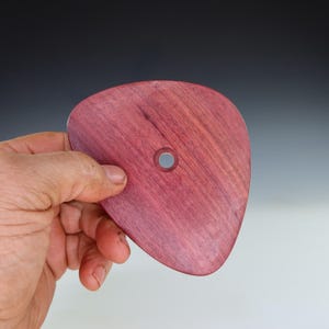 May include: A handcrafted, heart-shaped wooden pottery rib in a deep reddish-purple hue. The tool features a smooth, polished surface and a small, centered hole. The wood grain is visible, adding to its natural aesthetic. This pottery rib is designed for shaping and smoothing clay.