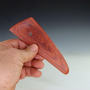 May include: A reddish-brown wooden pottery rib tool with a triangular shape and a small hole near the top. The tool is held in a hand, showcasing its smooth, polished surface and the natural wood grain.