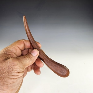 May include: A handcrafted, curved wooden tool with a smooth, dark brown finish. The tool has a rounded handle and a slender, pointed end. The wood grain is visible, adding to its natural aesthetic. The tool is held in a hand.