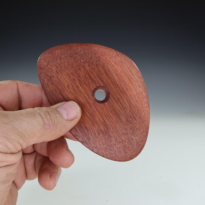 May include: A smooth, reddish-brown wooden sculpting tool with a hole in the center. The tool is shaped like an oval with rounded edges.