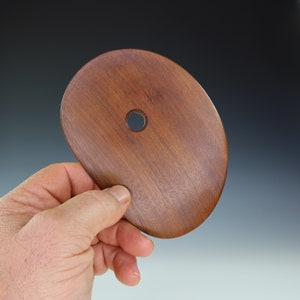 May include: A smooth, oval-shaped wooden tool with a hole in the center. The tool is made of dark brown wood and has a natural grain pattern.