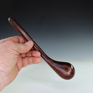 May include: A dark brown wooden spoon with a long handle and a rounded bowl. The spoon is held in a hand, showing its size and shape. The spoon's wood grain is visible. The spoon is likely for cooking or serving.