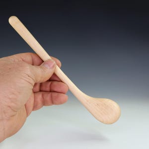 May include: A light-colored wooden spoon with a rounded bowl and a long, slender handle. The spoon is held in a hand, showcasing its natural wood grain and smooth finish. The spoon is approximately 10 inches long.