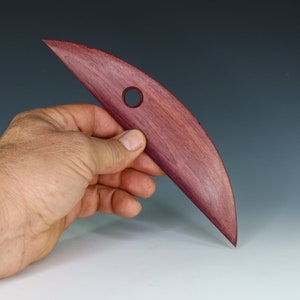 May include: A wooden sculpting tool with a curved blade and a hole in the handle. The tool is made of a dark reddish-purple wood.
