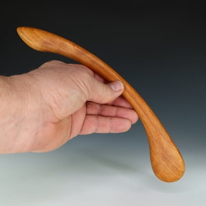 May include: A smooth, curved wooden spoon with a rounded tip. The spoon is made of light brown wood and is held in a hand.