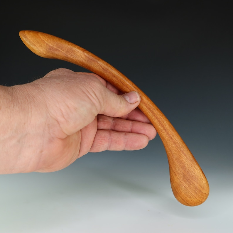 May include: A smooth, curved wooden spoon with a rounded tip. The spoon is made of light brown wood and is held in a hand.