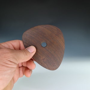 May include: A dark brown, wooden pottery rib tool with a rounded triangular shape and a small hole. The tool is held in a person's hand against a gradient background of gray and white.