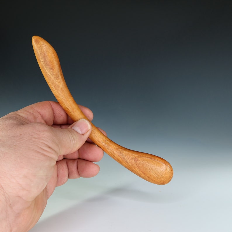 May include: A wooden tool with a curved handle and a rounded end. The tool is made of light brown wood and is held in a person's hand.