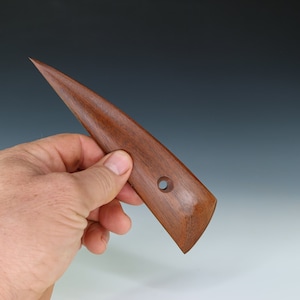 May include: A wooden tool with a pointed tip and a rounded handle. The tool has a hole in the handle. It is made of dark brown wood.