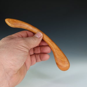 May include: A smooth, wooden massage tool with a curved handle and a rounded end. The tool is made of light brown wood and is held in a hand.