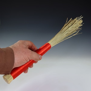 May include: A red and natural fiber brush with a long handle. The brush is held in a hand.