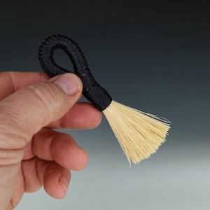 May include: A small black and natural bristle brush with a looped handle. The brush is held in a hand.