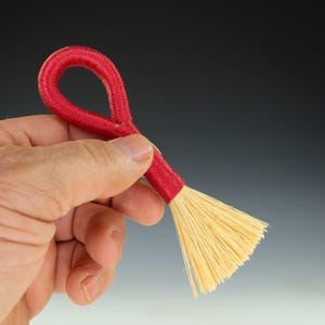 May include: A small red and natural fiber brush with a looped handle. The brush is held in a hand.