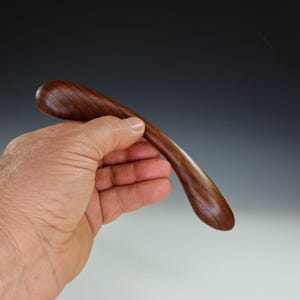 May include: A curved, wooden object with a smooth, polished finish. The wood grain is visible, showcasing a rich, dark brown color. The object is held in a hand, suggesting its size and shape. The design appears ergonomic, possibly a handle or decorative piece.