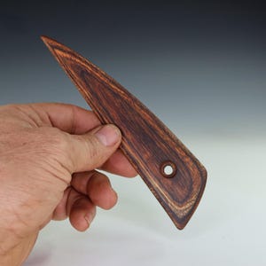 May include: A handcrafted wooden pottery rib with a dark brown grain pattern. The tool has a triangular shape with a rounded end and a hole. The wood grain is visible throughout the tool.