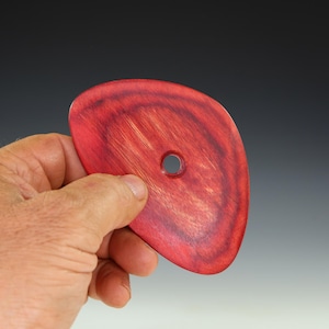 May include: A red wooden pottery rib tool with a hole. The tool is held in a hand and has a curved shape. The wood grain is visible, and the color is a deep, rich red.