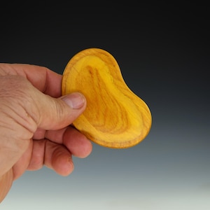 May include: A hand holding a smooth, yellow wooden object with a bean-like shape. The wood grain is visible, with darker brown lines contrasting against the yellow. The object appears to be a massage tool or a decorative item.
