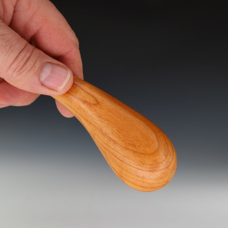 May include: A smooth, wooden spoon with a teardrop shape, crafted from cherry wood. The spoon has a natural wood finish and is held in a hand.