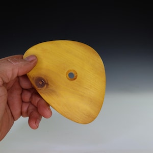 May include: A handcrafted, yellow-toned wooden pottery rib with a smooth, curved shape and a central hole. The wood grain is visible, and the rib is held by a hand against a gradient background.