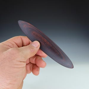 May include: A long, thin, oval-shaped wooden tool with a dark reddish-brown color. The wood has a natural grain pattern.