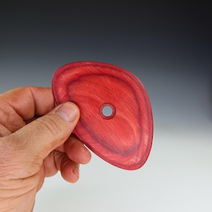 May include: A red wooden disc with a hole in the center. The disc is irregularly shaped and has a smooth, polished surface.