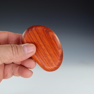 May include: A smooth, oval-shaped wooden coaster with a rich reddish-brown color and a wood grain pattern.